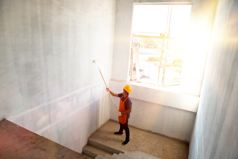 Room paint job. Asian white room painter with a paint roller in a great hall on a construction site.